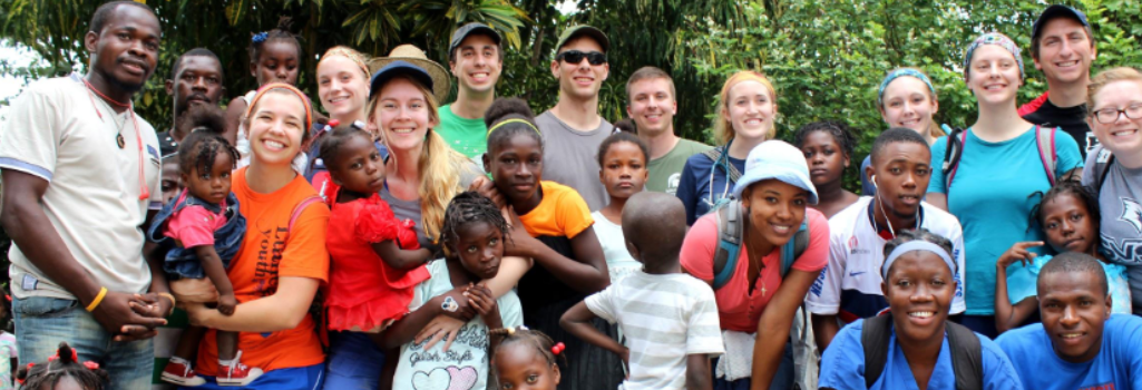 A large group of people stand smiling for a picture with green foliage behind them.